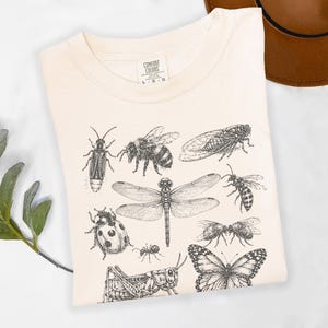 Pode incluir: Camiseta creme com uma ilustração detalhada em preto e branco de vários insetos, incluindo uma libélula, uma abelha e uma borboleta. A camiseta tem uma etiqueta que diz "Comfort Colors". Um chapéu marrom é visível no fundo.