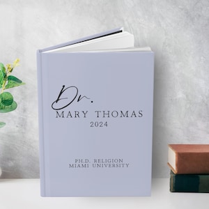 Puede incluir: Un libro de tapa dura de color lila claro con el texto "Dr. Mary Thomas 2024" y "Ph.D. Religion Miami University" en la portada.