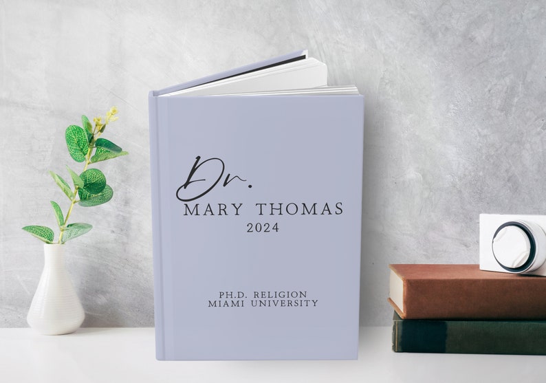Puede incluir: Un libro de tapa dura de color lila claro con el texto "Dr. Mary Thomas 2024" y "Ph.D. Religion Miami University" en la portada.