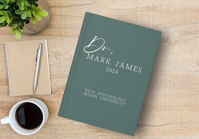 Puede incluir: Un diario de tapa dura verde azulado con el texto "Dr. Mark James 2024" y "Ph.D. Psychology Miami University" en letras blancas.