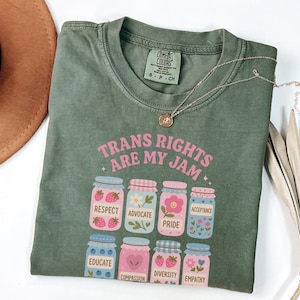 May include: Green t-shirt with the text "Trans Rights Are My Jam" in pink letters. The shirt features a graphic of jars with different words on them, including "Respect", "Advocate", "Pride", "Acceptance", "Educate", "Compassion", "Diversity", and "Empathy".