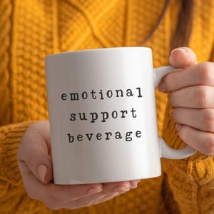 May include: A white ceramic mug with the words "emotional support beverage" printed in black. The mug is held by a person wearing a yellow knitted jumper.