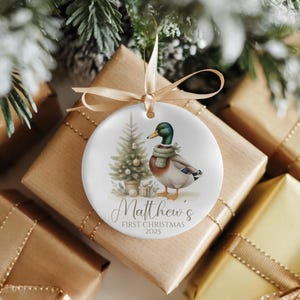 Könnte beinhalten: Ein runder, weißer Keramik-Weihnachtsbaumschmuck mit einem Stockenten-Motiv, die einen grünen Schal trägt, neben einem geschmückten Weihnachtsbaum. Der Text lautet Matthew's FIRST CHRISTMAS 2025. Der Anhänger ist mit einem dünnen Goldband versehen.