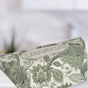 K&ouml;nnte beinhalten: Eine Kosmetiktasche mit gr&uuml;nem und wei&szlig;em Paisley-Muster. Die Tasche hat einen goldenen Rei&szlig;verschluss und den Namen "LAURIE" in einem dekorativen Rahmen. Die Tasche ist aus einem weichen, gemusterten Stoff.