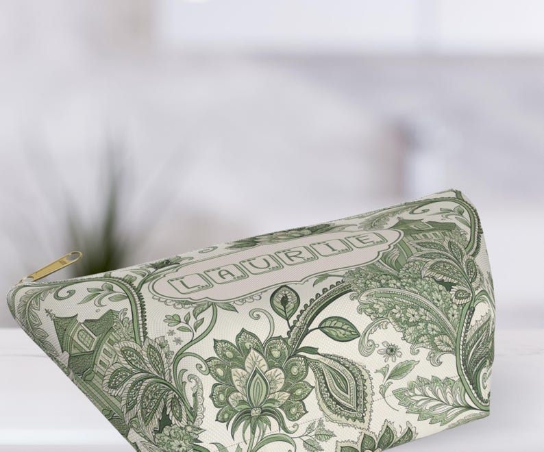 K&ouml;nnte beinhalten: Eine Kosmetiktasche mit gr&uuml;nem und wei&szlig;em Paisley-Muster. Die Tasche hat einen goldenen Rei&szlig;verschluss und den Namen "LAURIE" in einem dekorativen Rahmen. Die Tasche ist aus einem weichen, gemusterten Stoff.
