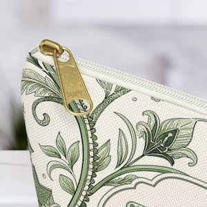 K&ouml;nnte beinhalten: Nahaufnahme einer Kosmetiktasche mit goldenem Rei&szlig;verschluss. Die Tasche hat einen cremefarbenen Hintergrund mit einem gr&uuml;nen Blumenmuster. Der Rei&szlig;verschlussanh&auml;nger ist mit den Buchstaben "YKK" versehen.
