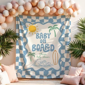 May include: A baby shower backdrop with a blue and white checkered pattern, featuring the text "Baby on Board" with a sun, palm trees, a surfboard, and waves. The scene is decorated with balloons and pillows.