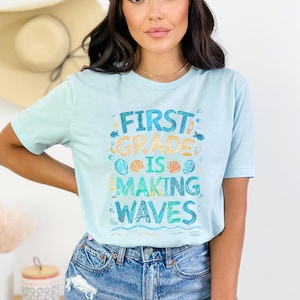 Pode incluir: Uma camiseta azul claro com o texto "First Grade is Making Waves" em um estilo colorido e desenhado &agrave; m&atilde;o. O texto &eacute; cercado por imagens de conchas, peixes e ondas.