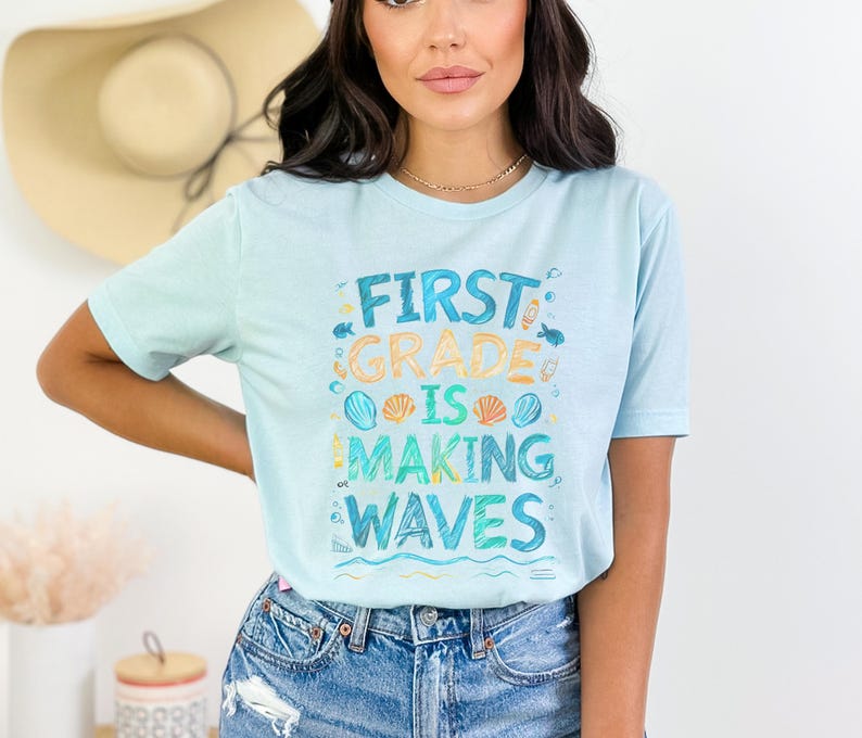 Pode incluir: Uma camiseta azul claro com o texto "First Grade is Making Waves" em um estilo colorido e desenhado &agrave; m&atilde;o. O texto &eacute; cercado por imagens de conchas, peixes e ondas.