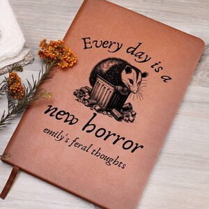 May include: A brown leather journal with the text "Every day is a new horror, emily's feral thoughts" and a black and white illustration of an opossum in a trash can.