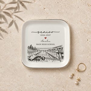 May include: A square white ceramic trinket dish with gold trim featuring a black ink illustration of a swimming pool with a swim cap and goggles. Text reads: senior 2026-2027, a red heart, Amelia, and MAIN HIGH SCHOOL in black lettering.