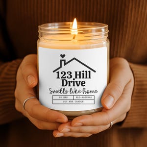 May include: A lit soy wax candle in a clear glass jar. The label features a house graphic with the text "123 Hill Drive Smells like home". The candle is held by a person wearing rings.