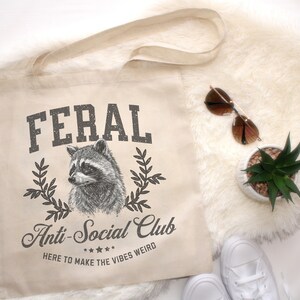 May include: A beige canvas tote bag with black text that reads "FERAL Anti-Social Club HERE TO MAKE THE VIBES WEIRD" and a black and white illustration of a raccoon.