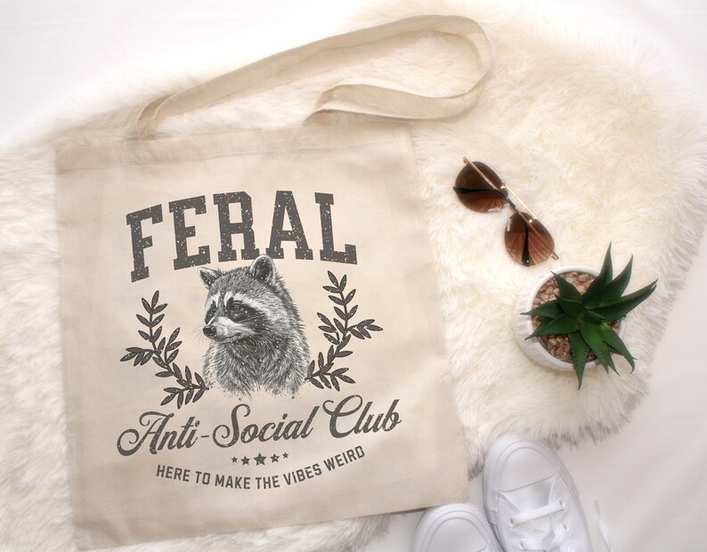 May include: A beige canvas tote bag with black text that reads "FERAL Anti-Social Club HERE TO MAKE THE VIBES WEIRD" and a black and white illustration of a raccoon.