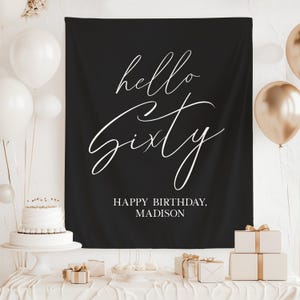 Pode incluir: Uma faixa preta com texto branco em itálico que diz "hello Sixty" e "HAPPY BIRTHDAY, MADISON". A faixa faz parte de uma configuração de festa de aniversário com balões, um bolo e presentes embrulhados.