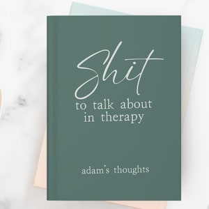 Op de afbeelding: Een teal-kleurig notitieboek met de woorden "Shit to talk about in therapy" in witte letters. De zin "adam's thoughts" staat onderaan gedrukt. Het notitieboek ligt op een wit marmeren oppervlak met een hartvormige schaal met gouden paperclips.