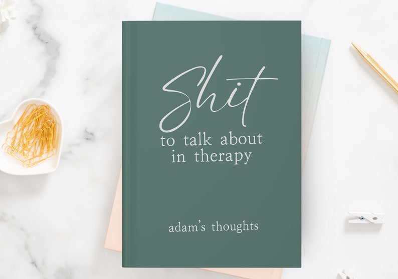 Op de afbeelding: Een teal-kleurig notitieboek met de woorden "Shit to talk about in therapy" in witte letters. De zin "adam's thoughts" staat onderaan gedrukt. Het notitieboek ligt op een wit marmeren oppervlak met een hartvormige schaal met gouden paperclips.