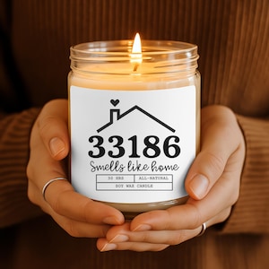 Op de afbeelding: Een brandende sojawas kaars in een heldere glazen pot, vastgehouden door handen. Het etiket luidt "33186 Smells like home" met een huisafbeelding en de tekst "30 HRS ALL-NATURAL SOY WAX CANDLE". De kaars heeft een warme, uitnodigende gloed.