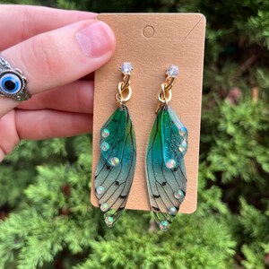 May include: A pair of green and blue fairy wing earrings with gold chain accents and clear rhinestones.