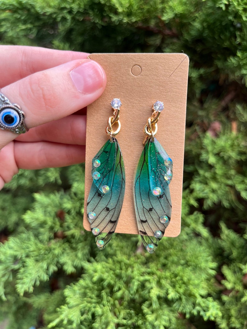 May include: A pair of green and blue fairy wing earrings with gold chain accents and clear rhinestones.