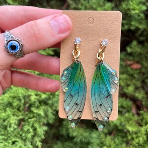 May include: A pair of green and blue iridescent fairy wing earrings with gold chain accents and clear rhinestones.