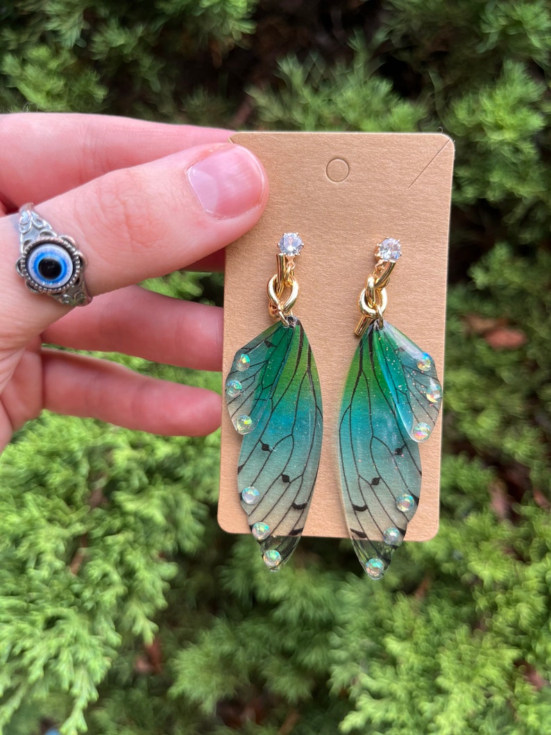 May include: A pair of green and blue iridescent fairy wing earrings with gold chain accents and clear rhinestones.