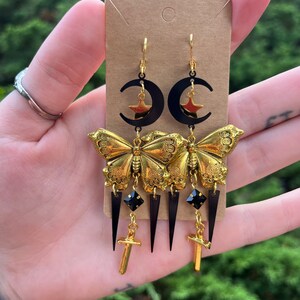 May include: Gold butterfly earrings with black crescent moons, stars, and sword charms. The earrings also feature black teardrop-shaped crystals.
