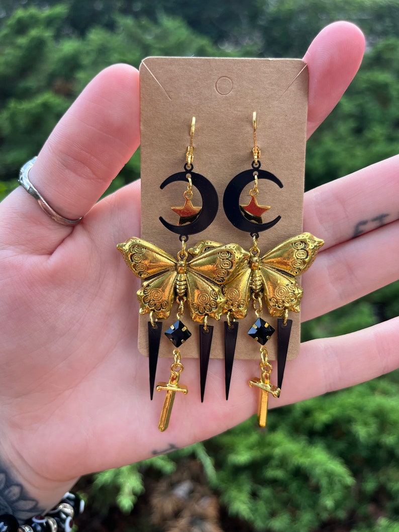 May include: Gold butterfly earrings with black crescent moons, stars, and sword charms. The earrings also feature black teardrop-shaped crystals.