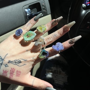 May include: A hand wearing several rings with different colored agate stones. The rings are set in silver and have a variety of shapes and sizes. The hand is also wearing a gray nail polish.