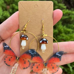 May include: Gold hoop earrings with a mushroom charm and a butterfly wing charm with a clear crystal hanging from the bottom.