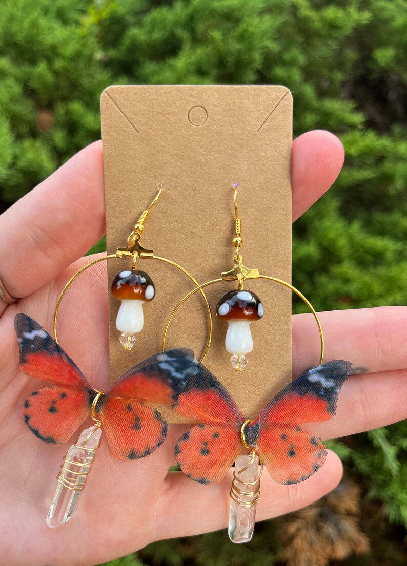 May include: Gold hoop earrings with a mushroom charm and a butterfly wing charm with a clear crystal hanging from the bottom.