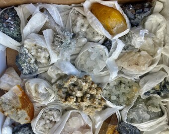 May include: A box filled with various raw crystals and minerals, wrapped in white tissue paper. The crystals vary in color, including clear, yellow, gray, and brown. Some are clustered, while others are individual specimens.