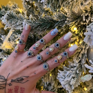 May include: A hand wearing multiple silver rings with blue evil eye charms and white pearl accents. The rings are worn on multiple fingers. The hand is in front of a snowy Christmas tree.