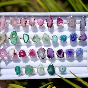 May include: A white box with a variety of agate rings. The rings are in different colors, including pink, green, blue, and purple. The rings are arranged in rows in the box.