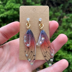 May include: A pair of dangle earrings featuring translucent wings with a gradient of pink, purple, and black. The wings have a musical note pattern and are accented with clear rhinestones.