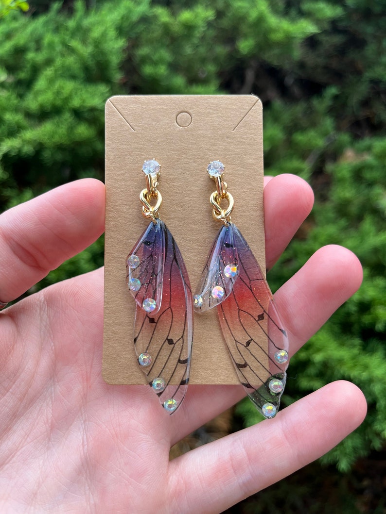 May include: A pair of dangle earrings featuring translucent wings with a gradient of pink, purple, and black. The wings have a musical note pattern and are accented with clear rhinestones.