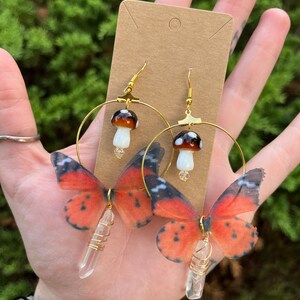 May include: Gold hoop earrings with a clear crystal charm and a butterfly wing design. The butterfly wings are orange and black with white spots. The earrings also feature a small mushroom charm.