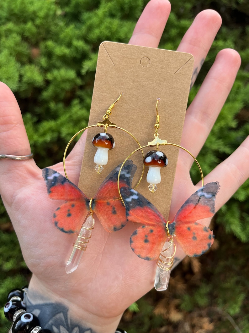 May include: Gold hoop earrings with a clear crystal charm and a butterfly wing design. The butterfly wings are orange and black with white spots. The earrings also feature a small mushroom charm.