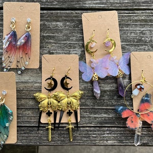 May include: Six pairs of handmade earrings featuring various designs, including butterfly wings, moons, stars, crystals, and mushrooms. The earrings are displayed on brown card stock.