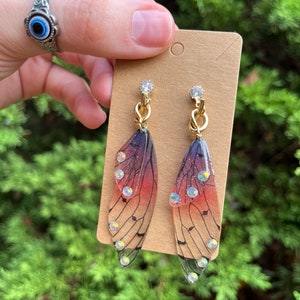 May include: A pair of gold earrings with a butterfly wing design. The wings are translucent and have a gradient of pink, purple, and black. The wings are decorated with small, clear rhinestones.