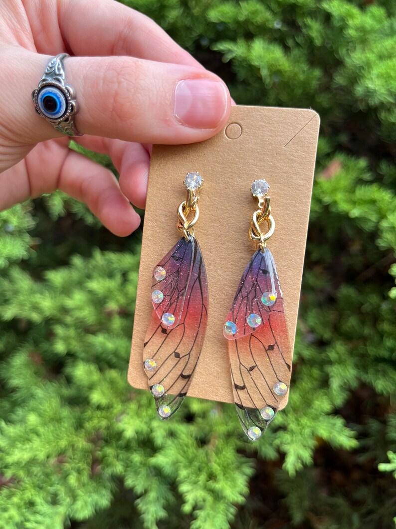 May include: A pair of gold earrings with a butterfly wing design. The wings are translucent and have a gradient of pink, purple, and black. The wings are decorated with small, clear rhinestones.