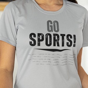May include: A woman wearing a gray t-shirt with the text "GO SPORTS!" printed on it. She is holding a wooden tray with a bowl of guacamole and chips, a bowl of chips, and a bowl of celery sticks.