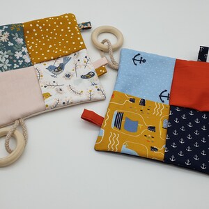 Peut inclure: Deux jouets sensoriels pour bébé faits à la main avec différents motifs de tissu. Un jouet est bleu, jaune, rouge et bleu marine avec un motif d'ancre. L'autre jouet est rose, vert, jaune et blanc avec un motif d'oiseau. Les deux jouets ont des anneaux en bois attachés.