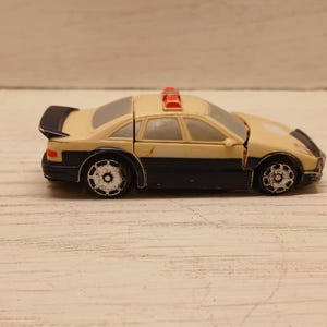 Puede incluir: Un coche de policía de juguete crema y azul marino con alerón negro y luces rojas. El coche tiene ruedas plateadas y está sobre una superficie clara. El coche mide unos 12 cm de largo.