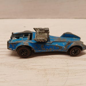 Op de afbeelding: Een vintage blauwe speelgoedauto met een grote motor bovenop. De auto heeft rode wielen en vertoont tekenen van slijtage, met afbladderende verf. De speelgoedauto is een verzamelobject.