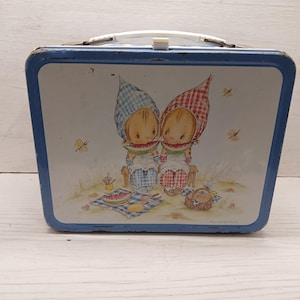 May include: A rectangular blue metal lunch box featuring a white center panel with an illustration of two children wearing checkered hats while eating watermelon slices on a picnic blanket, complete with a white plastic handle and metal latch.