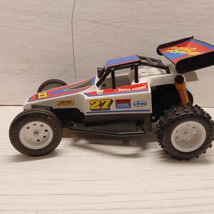 May include: A white, black, blue, and red toy racing buggy with the number 27 on the side. The vehicle features a rear spoiler labeled ROAD HOPPER, white wheels with black tires, a roll cage, and various decals including SUPER RACING and Arai.
