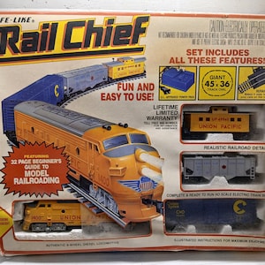 Op de afbeelding: Een vintage Life-Like Rail Chief elektrische treinset. De doos toont een oranje en blauwe diesellocomotief, wagons en rails. De set bevat een 114 x 91 cm lange ovale baan en een beginnersgids voor modelspoorwegen.