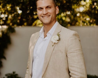 May include: A man wearing a light brown linen suit jacket with a white button-down shirt and a white flower boutonniere. He is smiling and looking at the camera.