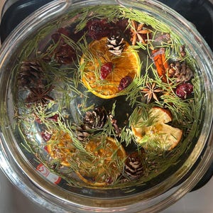 May include: A glass bowl filled with water, orange slices, pine needles, cranberries, star anise, pine cones, and cinnamon sticks. The arrangement creates a festive and aromatic display, ideal for home fragrance.